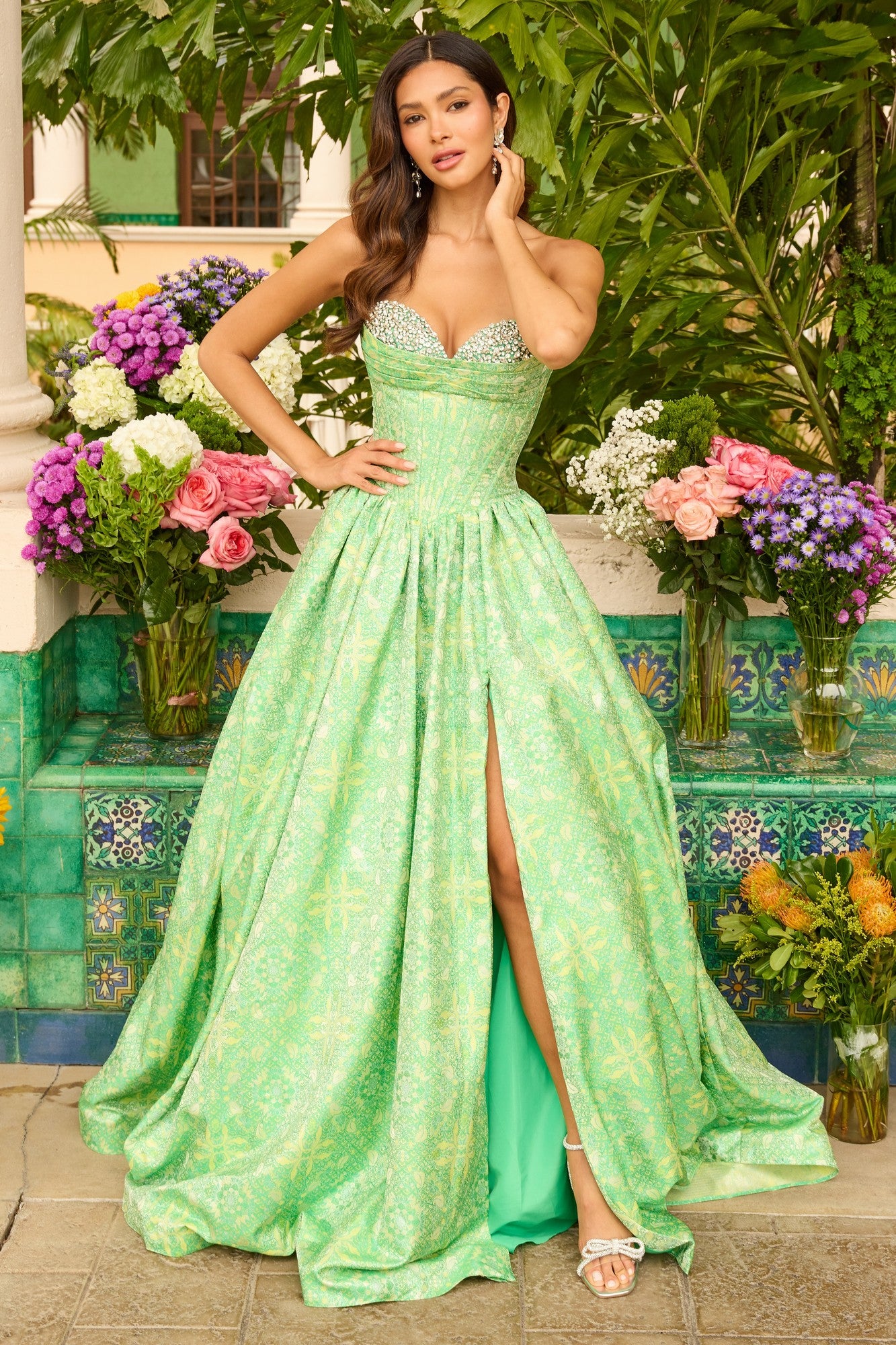 A woman stands outdoors in the green Ava Presley 42318 long prom dress, a strapless gown with a high slit and floral print, surrounded by colorful flowers and lush greenery.